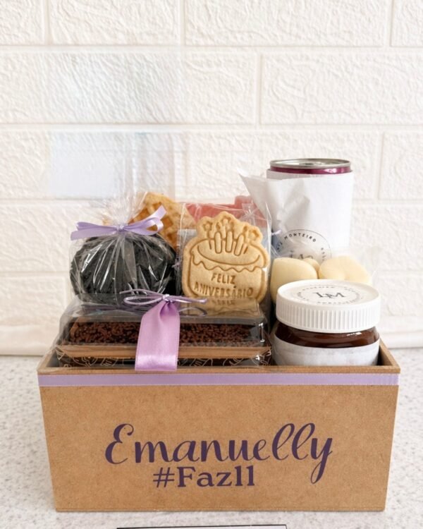 Cesta de café da manhã infantil Box Kids em caixa de MDF personalizada com o nome Emanuelly. A cesta contém bolo de chocolate com fita lilás, biscoito artesanal escrito Feliz Aniversário, pote de creme de avelã, waffle e doces embalados.