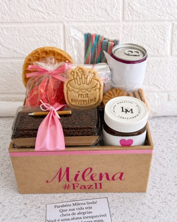 Presente de aniversário infantil Box Kids em caixa de MDF decorada em tons de rosa com o nome Milena. A cesta inclui bolo de chocolate, biscoito decorado de Feliz Aniversário, balas coloridas tipo Fini, creme de avelã, doces e um cartão de felicitações