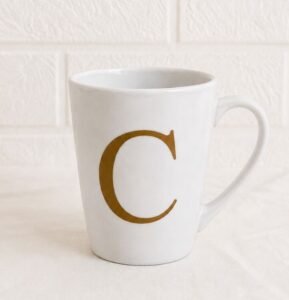 Caneca branca de cerâmica personalizada com a inicial C em dourado. Item exclusivo para compor e personalizar cestas de café da manhã.