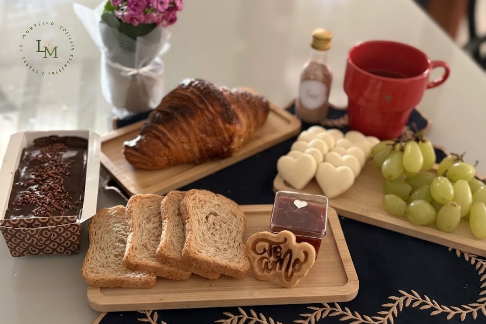 Mesa posta com itens da entrega de cesta de café da manhã em Ibirité: croissant, pães, geleia, bolo, uvas e caneca vermelha